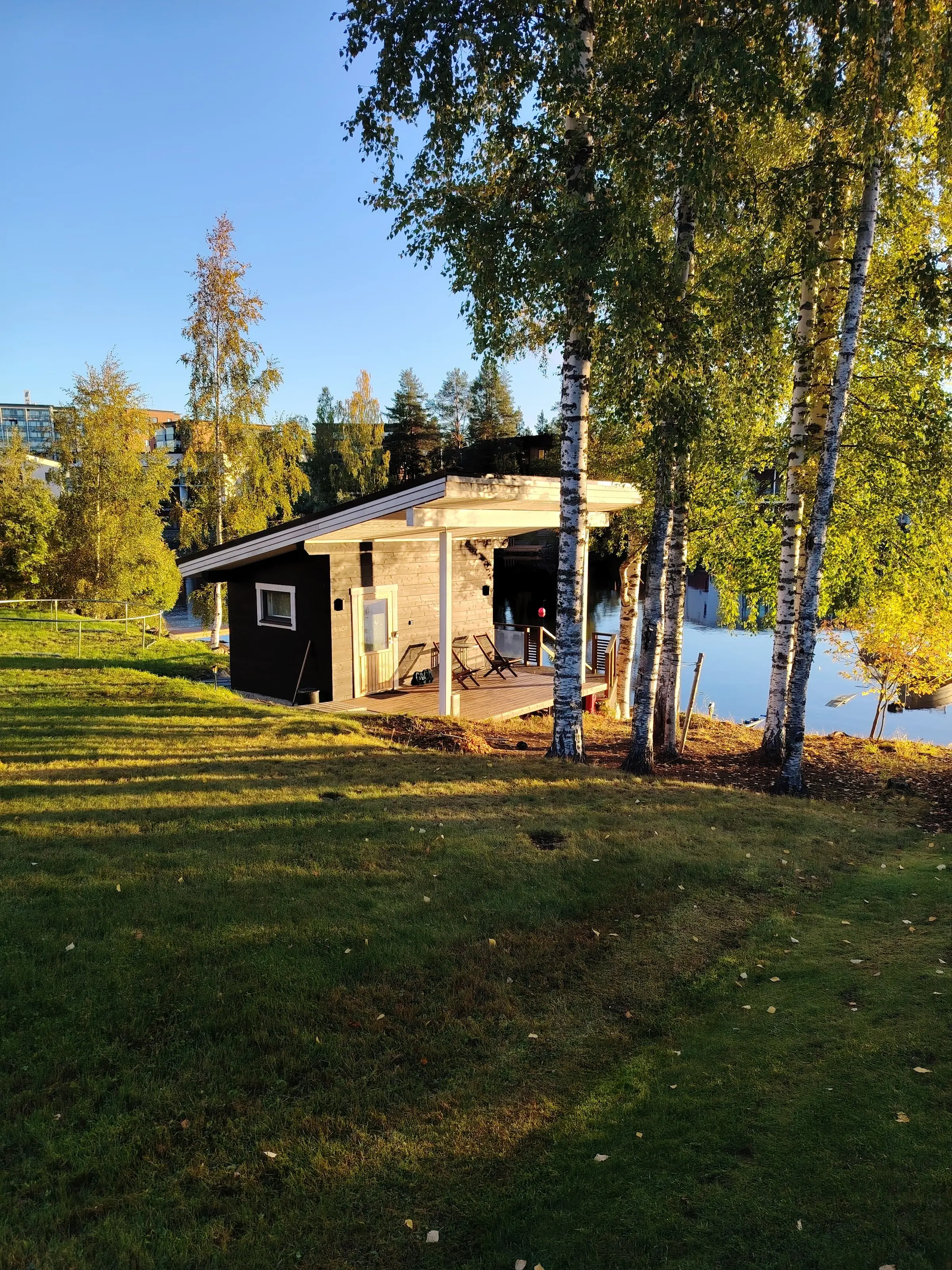 Rantasauna on the lakeshore, seen across the lawn, with birch trees and the lake on the right.